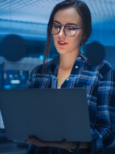 Portrait of a Smart Seductive Young Woman Wearing Glasses Holds Laptop. In the Background Technical Department Office with Specialists Working and Functional Data Server Racks