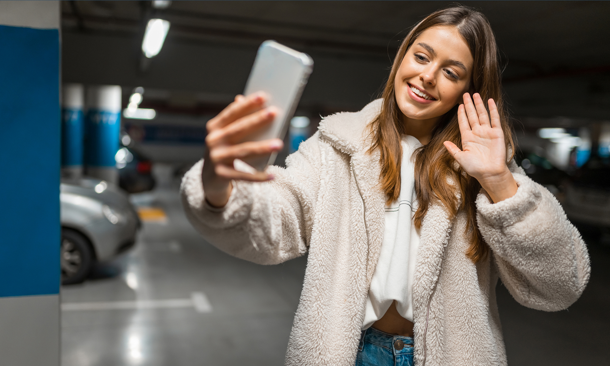 Beautiful girl takes selfie in the underground parking. Fashionable young woman with smartphone talking on video call and waving into camera