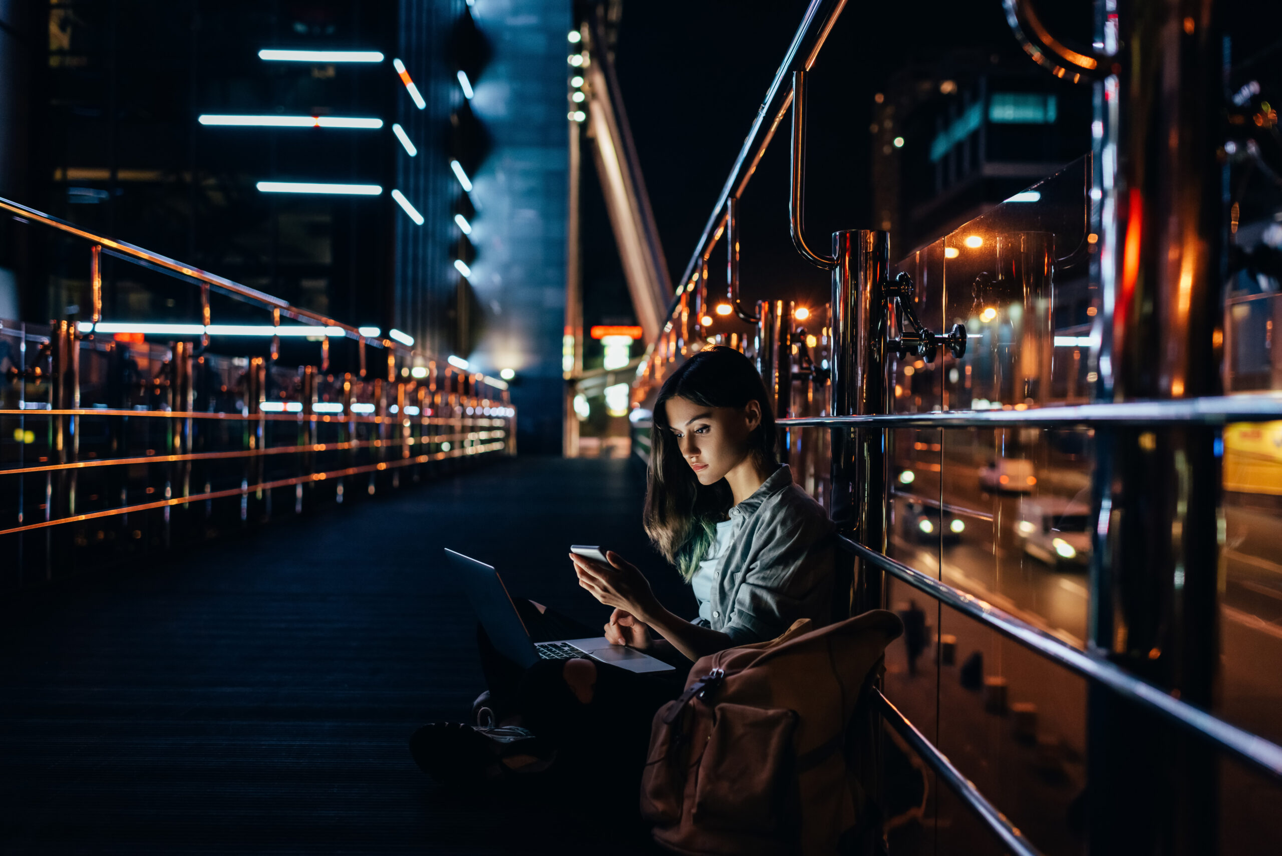 side view of young woman with laptop on knees using smartphone with night city on background