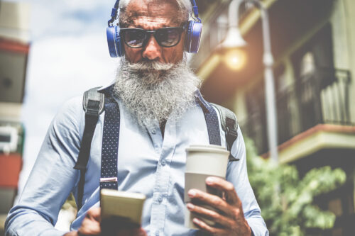 Happy senior man listening music playlist app and drinking coffee at early morning time - Technology, trendy lifestyle, and joyful elderly lifestyle concept - Focus on headphones