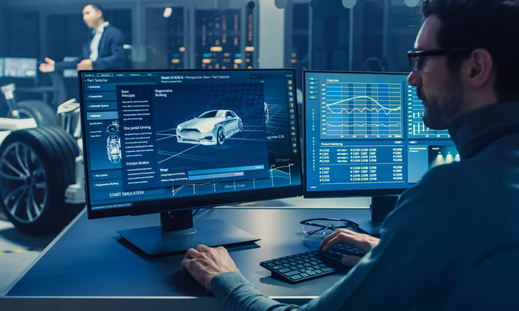 Professional Engineer Works on a Computer with a 3D CAD Software and Tests the Electric Car Chassis Prototype with Wheels, Batteries and Engine Standing in a High Tech Development Laboratory.